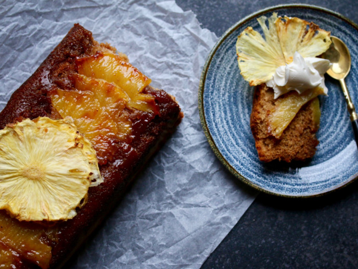 Pineapple upside down cake on butter paper next to a plate with a slice of the same with a pineapple piece and cream