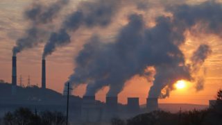 A distant view of industrial chimneys with smoke coming out against a setting sun.