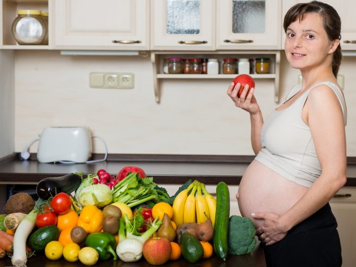 A pregnant woman holding an apple standing in front of a table full of vegetables and fruits.