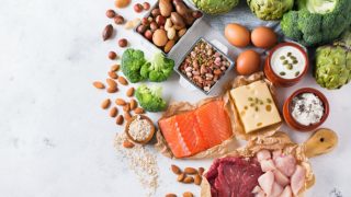 Foods rich in protein (fish, beef, chicken, almonds, broccoli, cheese, yogurt, artichokes, lentils, quinoa, oat, and dry fruits) on a white wooden table