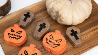 A whole pumpkin, and tinier cutouts of coffin and skulls on a wooden chopping board besides a candle
