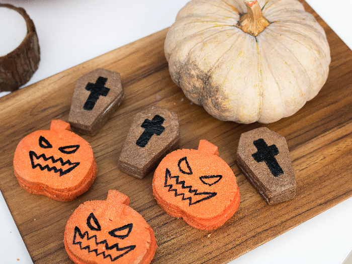 A whole pumpkin, and tinier cutouts of coffin and skulls on a wooden chopping board besides a candle