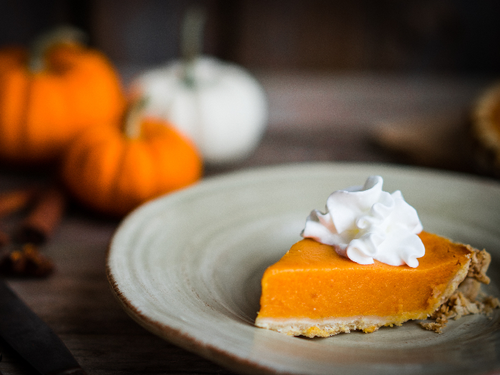 White and orange pumpkins and spices in the background of a slice of pumpkin pie topped with whipped cream