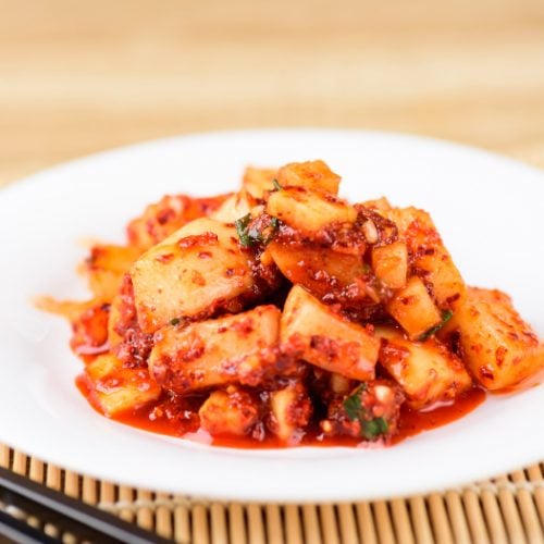 A close up shot of radish kimchi kept on a white plate with chopsticks