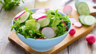 A white bowl with radish and cucumber salad on a wooden cutting board with sliced vegetables behind