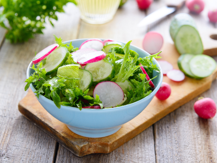 A white bowl with radish and cucumber salad on a wooden cutting board with sliced vegetables behind