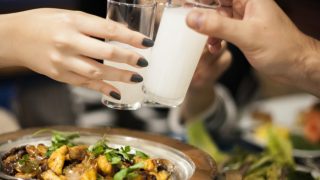 Two hands clinking shot glasses with white liquor over a bowl of food.