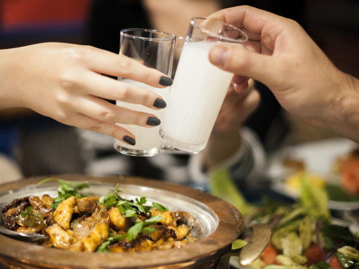 Two hands clinking shot glasses with white liquor over a bowl of food.