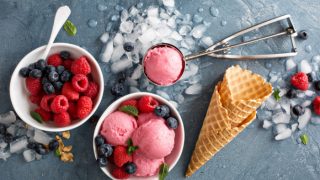 Flatline view of bowls of berries, ice-cream, cones, and a scoop of ice-cream over ice cubes on a slate surface.