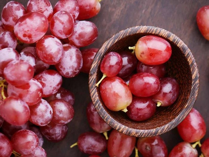 Red grapes kept in a wooden bowl next to a bunch of the fruit, placed atop a wooden table