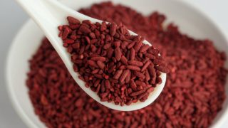 A close-up shot of a spoon full of red yeast rice against a hazy picture of a white bowl containing the same
