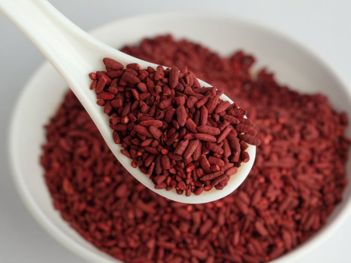 A close-up shot of a spoon full of red yeast rice against a hazy picture of a white bowl containing the same