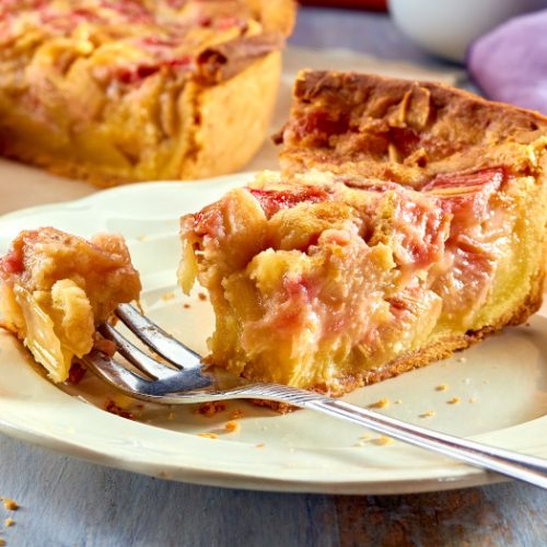 A slice of homemade rhubarb pie on a plate eaten with fork on a wooden table