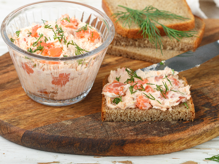 Salmon rillette in a bowl and spread on a bread slice with bread in the background