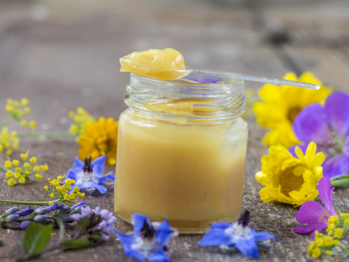A small bottle of organic royal jelly with a little spoon on it surrounded by different colorful flowers