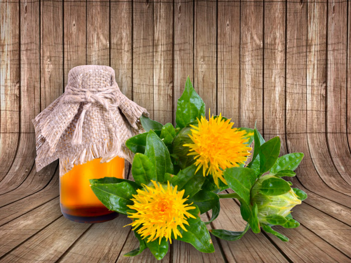 A jar of safflower oil covered with a jute cloth and safflower flowers on a wooden table