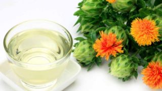 A glass of safflower oil kept next to safflowers against a white background