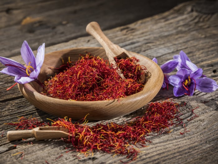 A wooden bowl and scoop filled with saffron and surrounded by fresh saffron flowers on a wooden table