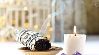 A lit sage smudge stick, next to a white candle on a white table with crystals