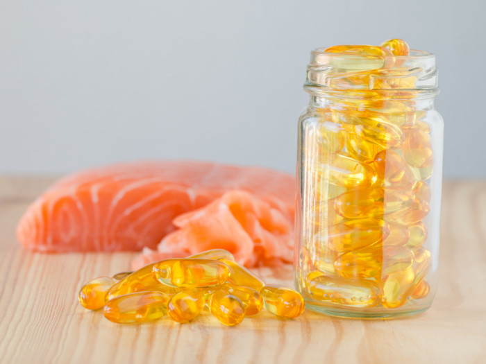 Close-up of glass bottle full of yellow pills, placed next to more pills on a wooden surface with a blurry fish piece at the back.