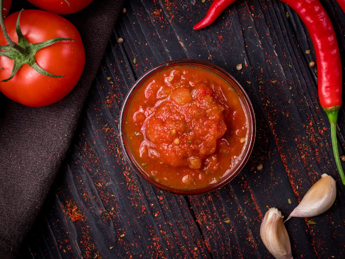A bowl full of salsa sauce kept atop a table next to garlic cloves, red chilies, and tomatoes