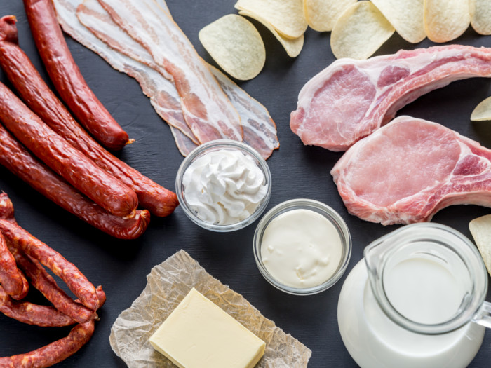 Flatline view of food high in fats, butter, sausages, bacon, dairy, chips, and meat chops on a slate background.