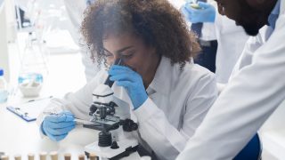 Female scientist looking through a microscope with a male and female colleague standing nearby