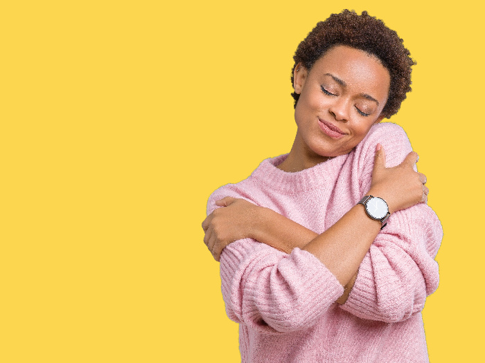 A woman hugging herself with a smile across her face against a yellow background