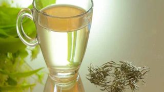 A cup of light-colored tea in a glass mug, placed along with tea leaves on a glass surface.