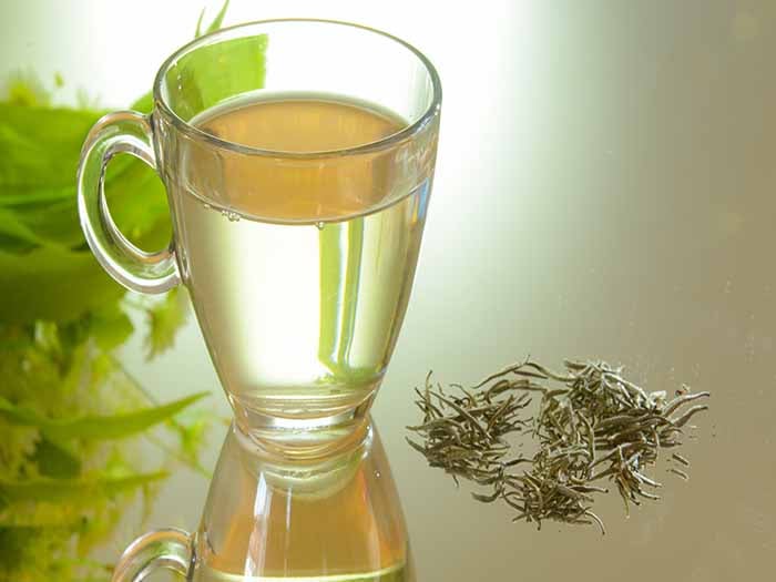 A cup of light-colored tea in a glass mug, placed along with tea leaves on a glass surface.