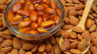 A bowl of soaked almonds with dried almonds on a wooden spoon and background