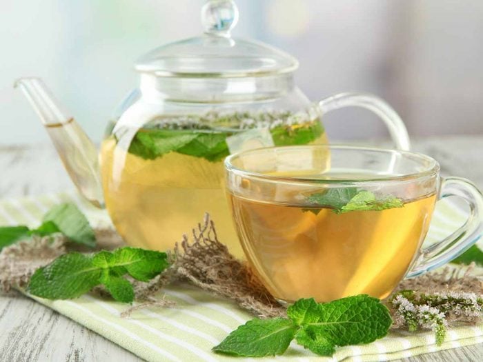 A kettle and cup of spearmint tea surrounded with fresh spearmint leaves on a cloth