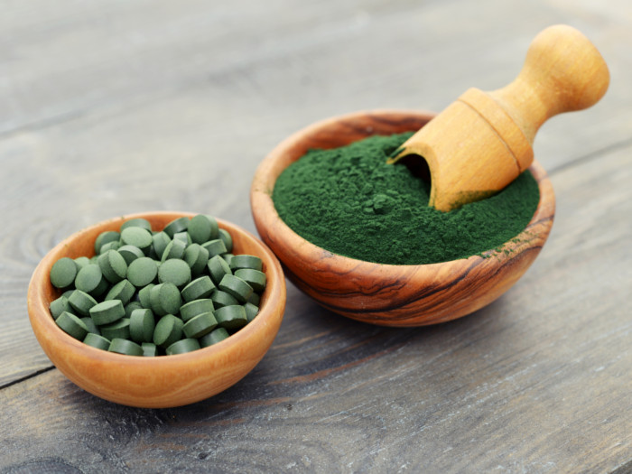 Wooden bowls of green spirulina powder and spirulina pills on a dark counter