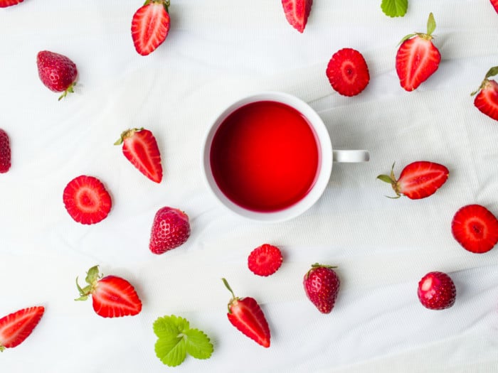 A cup of strawberry tea, whole and sliced strawberries on a white counter.