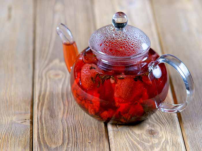 A glass teapot with cut strawberries and whole spices on a wooden counter