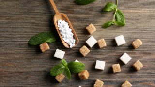 A wooden scoop filled with sugar tablets, brown and white sugar cubes with mint on a wooden table