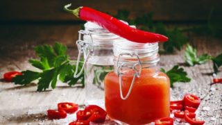 Close-up of two jars with sauce, a chili placed atop, chili pieces scattered around and coriander leaves at the back