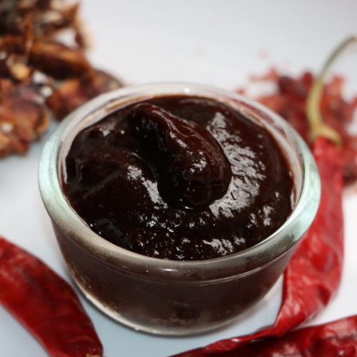 A small bowl of tamarind chutney on a white plate with scattered red chillies and tamarind