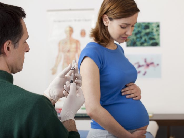 A pregnant woman looking serenely at her belly, while a doctor prepares a Tdap shot