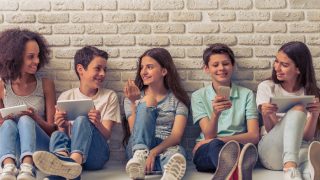 Group of teenage boys and girls is using gadgets, talking and smiling, sitting against white brick wall
