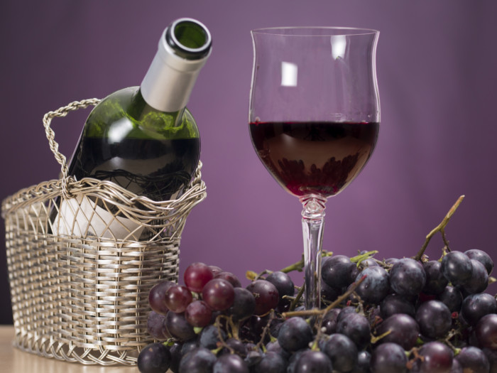 A glass of red wine placed inside bunches of grapes, alongside a basket containing a wine bottle.