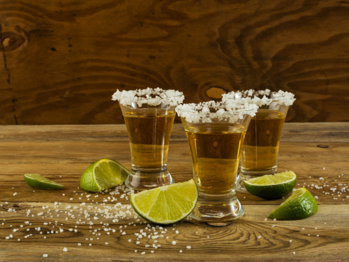 Three tequila glasses kept atop a wooden table with lemon slices cut and kept next to the glasses