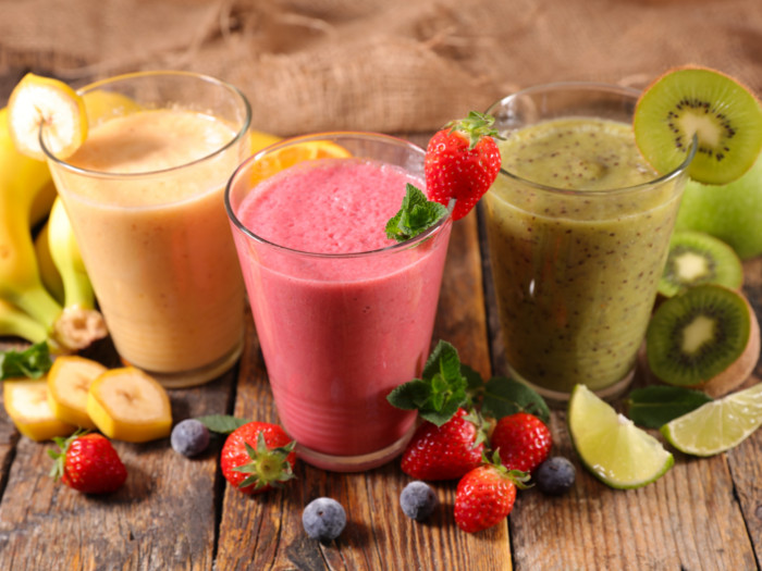 Glasses of banana, strawberry, and lime and kiwi smoothies with fruits scattered beneath