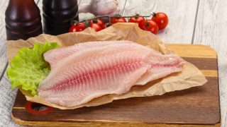 Tilapia fillets on parchment paper, with lettuce, cherry tomatoes, and garlic in the background