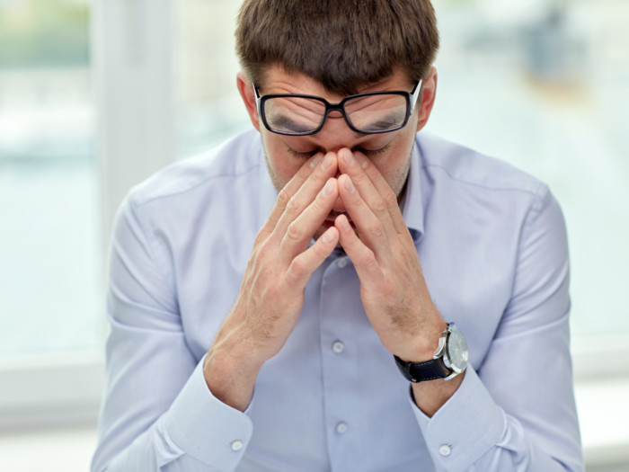 A close up of an upset man who is rubbing his eyes and has pushed up his reading glasses