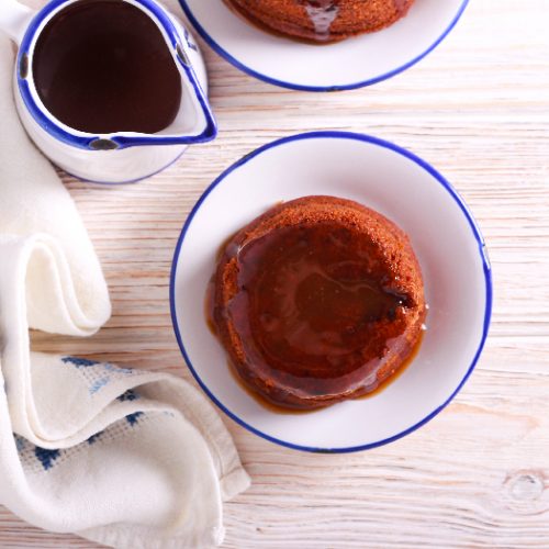 Top view of sticky toffee pudding in a white and blue bowl with a small jug of toffee sauce