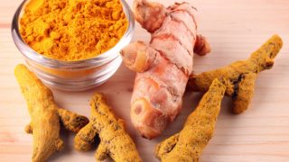 A bowl of turmeric powder and a couple of turmeric roots on a wooden table