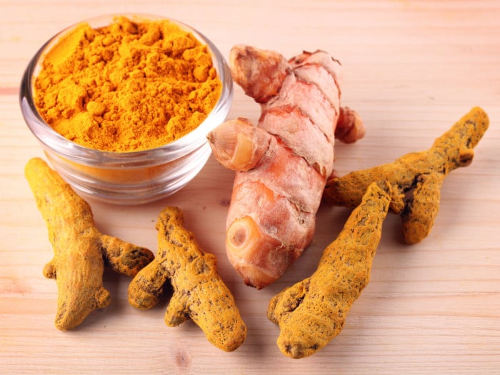 A bowl of turmeric powder and a couple of turmeric roots on a wooden table