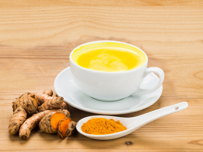 A small cup of turmeric beverage and turmeric roots with a spoonful of turmeric powder near it