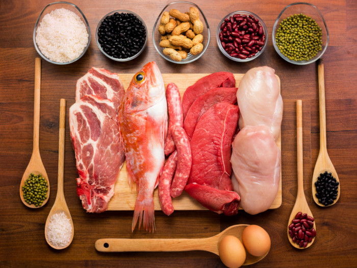 Different types of fresh raw meat (fish, pork, beef, chicken, and red meat) with uncooked proteins (legumes, nuts, rice, and eggs) on a wooden table